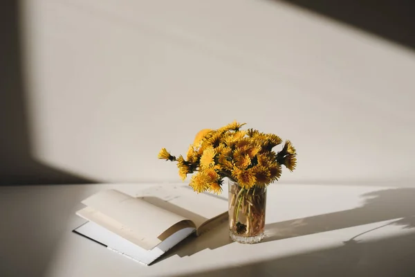 A book and a bunch of yellow dandelions.  Still life with wild flowers.  Play of light and shadow. Summer floral background