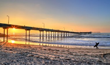 San Diego, Kaliforniya'da pier yakınındaki Ocean Beach üzerinde bir sörfçü yürür.