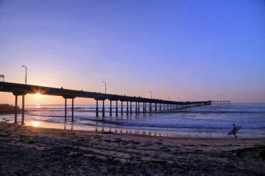 San Diego, Kaliforniya'da pier yakınındaki Ocean Beach üzerinde bir sörfçü yürür.