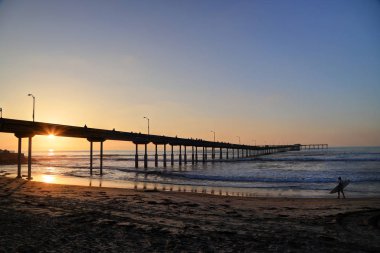 San Diego, Kaliforniya'da pier yakınındaki Ocean Beach üzerinde bir sörfçü yürür.