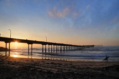 San Diego, Kaliforniya'da pier yakınındaki Ocean Beach üzerinde bir sörfçü yürür.