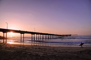 San Diego, Kaliforniya'da pier yakınındaki Ocean Beach üzerinde bir sörfçü yürür.
