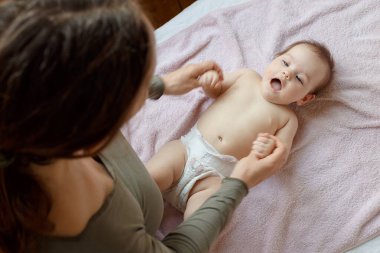 Anne ve çocuk beyaz bir yatakta. Bebek bezi takmış bir kız. Anne bebeğiyle oynuyor..