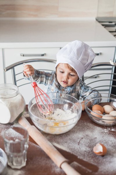 adorable little kid in chef hat preparing dough with whisk at kitchen