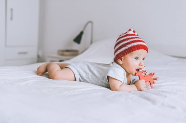 infant child in striped red and white hat biting wooden deer decoration in bed clipart