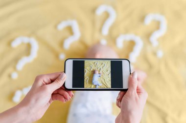 cropped shot of mother taking top view photo of child sleeping surrounded with question marks on yellow bed clipart