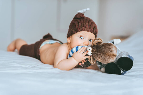 infant child in knitted deer costume playing with toy moose in bed