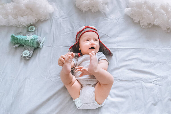 top view of dreamy infant child in pilot hat with toy plane surrounded with clouds made of cotton in bed