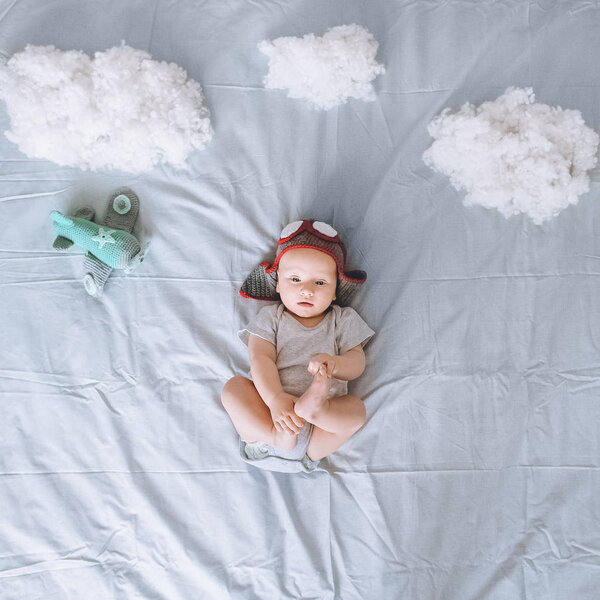 top view of dreamy infant child in knitted pilot hat with toy plane surrounded with clouds made of cotton in bed