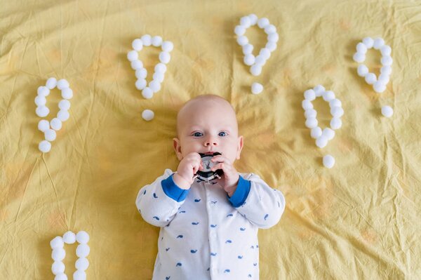top view of infant child surrounded with exclamation marks made of cotton balls biting alarm clock in morning in bed