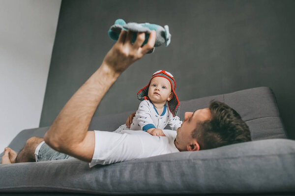 father holding toy plane while lying on sofa and playing with adorable infant child wearing knitted pilot hat