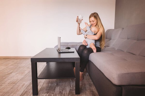 mother hugging infant child and holding glass of water while sitting on sofa and using laptop 