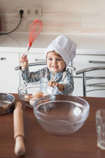 Happy little kid in chef hat with whisk and eggs in bowl — Stock Photo