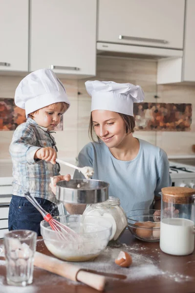 Madre y adorable niño en sombreros de chef preparando masa en la mesa desordenada en la cocina - foto de stock