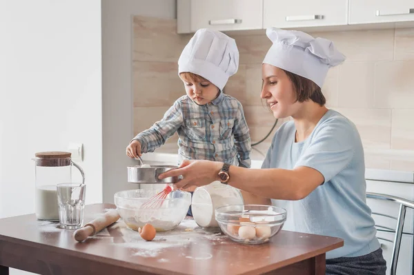 Madre joven y adorable niño en sombreros de chef preparando masa en la mesa desordenada en la cocina - foto de stock