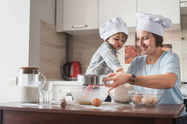 Glückliche junge Mutter und Kind in Kochmützen beim Teigzubereiten in der Küche — Stockfoto