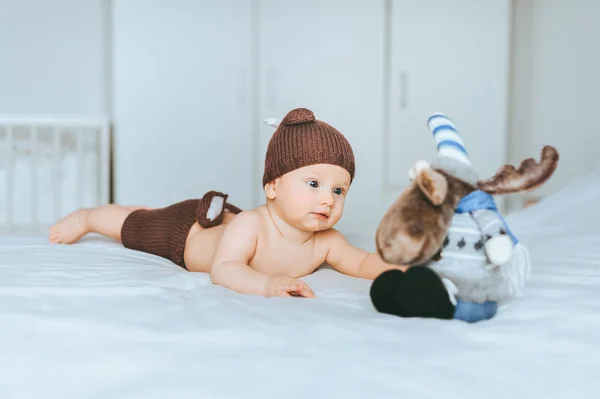 Niño pequeño en pantalones cortos de punto de ciervo y sombrero jugando con alce de juguete en la cama - foto de stock