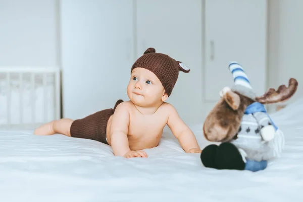 Adorable infant child in knitted deer shorts and hat in bed with toy moose — Stock Photo