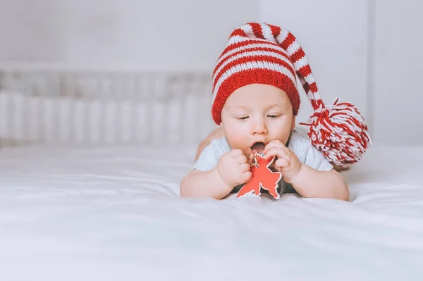 Infant child in red and white striped hat with pompom playing with toy angel in bed — Stock Photo