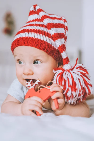 Close-up portrait of infant child in red and white striped hat with pompom playing with toy deer in bed — Stock Photo