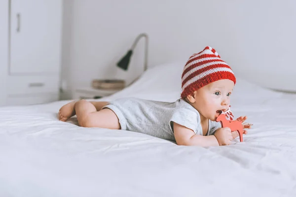 Infant child in striped red and white hat biting wooden deer decoration in bed — Stock Photo