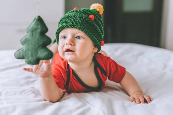 Infant child in adorable knitted hat in shape of christmas tree playing with holiday decoration in bed — Stock Photo