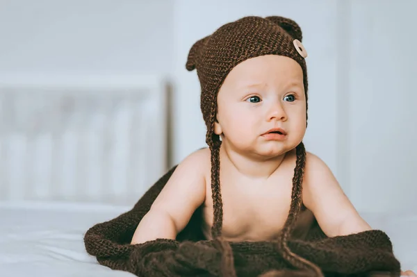 Niño pequeño en sombrero de punto marrón con manta en la cama mirando hacia otro lado - foto de stock