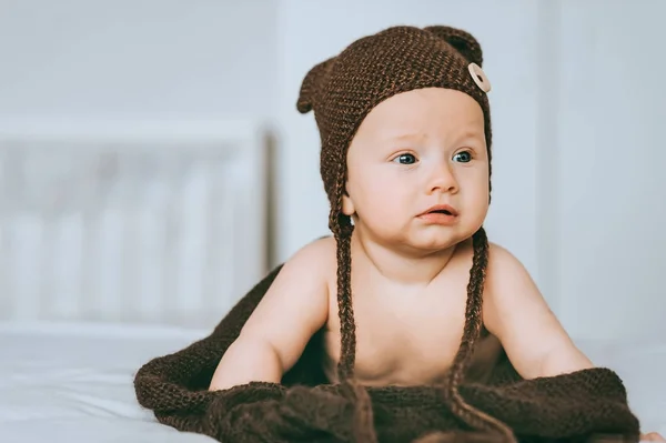 Infant child in brown knitted hat with blanket in bed looking away — Stock Photo
