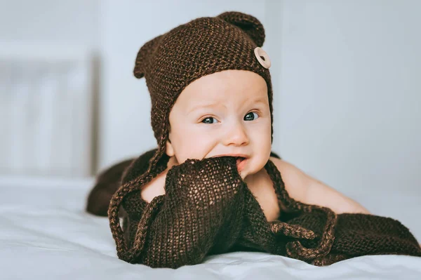 Infant child in brown knitted hat biting blanket in bed — Stock Photo