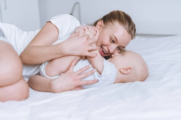 Joven feliz madre y su bebé bebé abrazándose en la cama - foto de stock