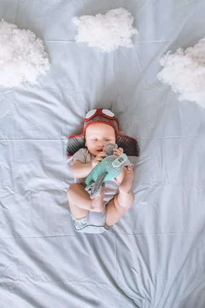 Vue de dessus du beau bébé enfant en chapeau de pilote avec avion jouet entouré de nuages en coton au lit — Photo de stock