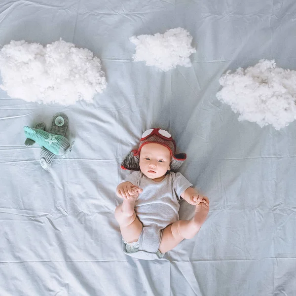 Vista superior da criança adorável infantil em chapéu piloto de malha com avião de brinquedo cercado por nuvens feitas de algodão na cama — Fotografia de Stock