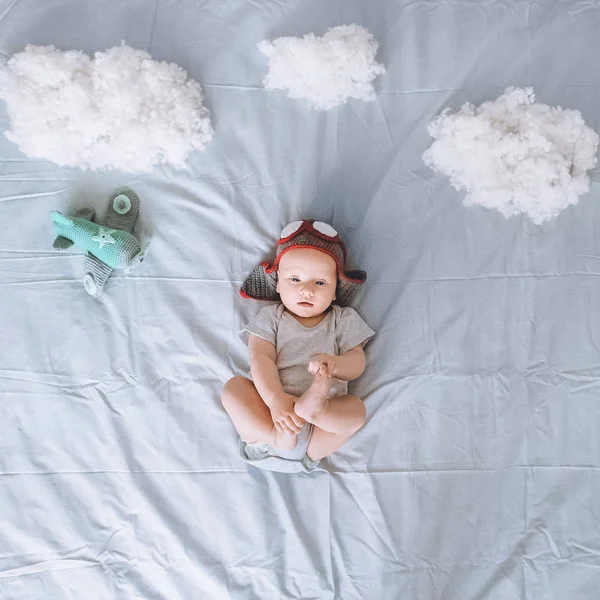 Vue de dessus du bébé rêveur en bonnet de pilote tricoté avec avion jouet entouré de nuages en coton au lit — Photo de stock