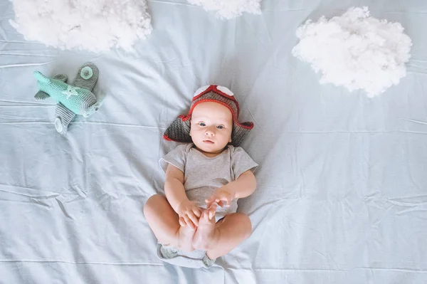 Vue de dessus de mignon enfant en bas âge en bonnet pilote tricoté avec avion jouet entouré de nuages en coton au lit — Photo de stock