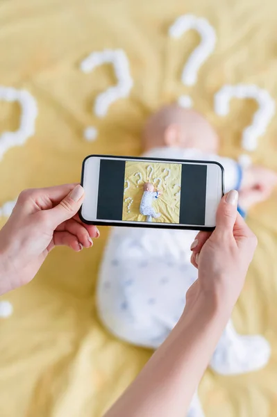 Fotografía recortada de la madre tomando la foto de vista superior con el teléfono inteligente del niño durmiendo en la cama rodeado de signos de interrogación - foto de stock