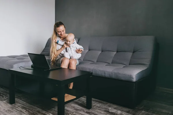 Joven madre y su hijo pequeño mirando a la computadora portátil mientras está sentado en el sofá en casa - foto de stock