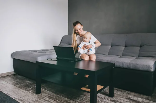 Sonriente joven madre e hijo lactante usando el ordenador portátil juntos mientras están sentados en el sofá en casa - foto de stock