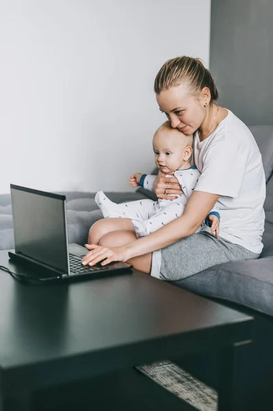 Junge Frau benutzt Laptop, während sie kleines Baby zu Hause trägt — Stockfoto