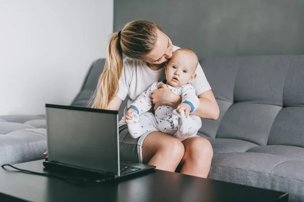 Beautiful young mother kissing adorable infant child while using laptop at home — Stock Photo