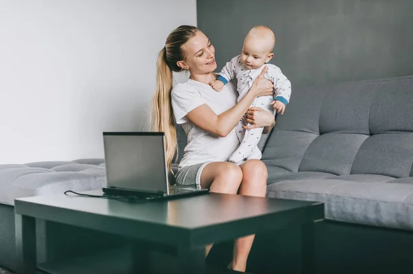 Feliz joven madre sosteniendo niño adorable mientras usa el ordenador portátil en casa - foto de stock