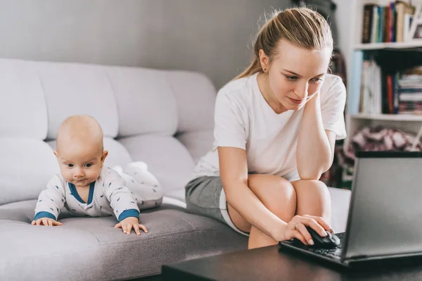 Madre joven usando el ordenador portátil, mientras que el niño pequeño acostado en el sofá - foto de stock