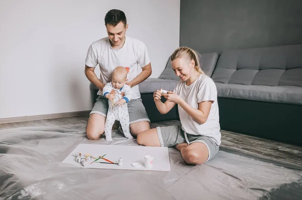 Familia joven y feliz con un niño pequeño pintando juntos en el suelo en casa - foto de stock