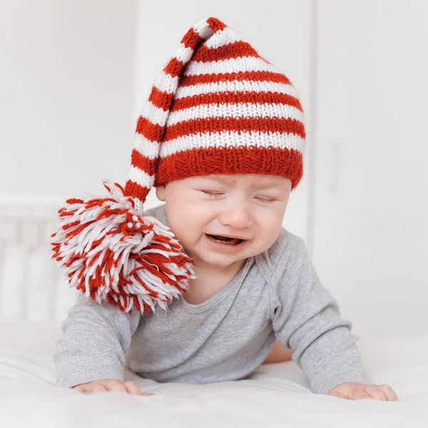 Portrait of little crying infant child in funny knitted hat lying on bed — Stock Photo