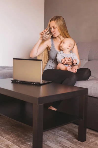 Junge Frau trinkt Wasser und hält niedliches kleines Kind, während sie auf dem Sofa sitzt und Laptop benutzt — Stockfoto