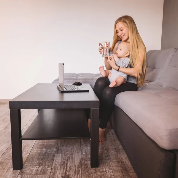 Vista lateral de la madre abrazando al bebé y sosteniendo un vaso de agua mientras está sentada en el sofá y usando la computadora portátil - foto de stock