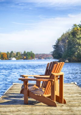 Mavi göldeki ahşap bir rıhtımda iki Muskoka sandalyesi 