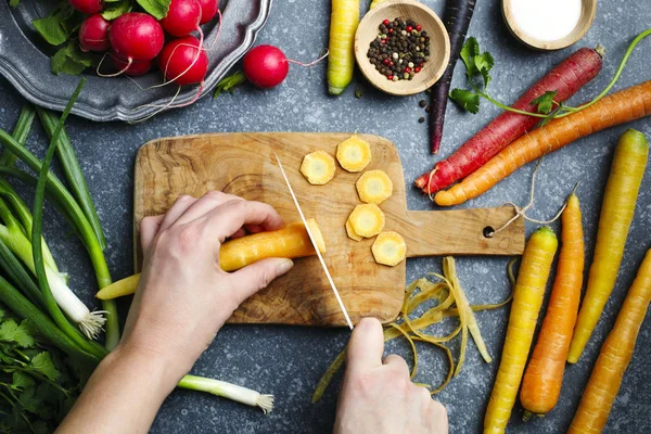 Manos de mujer cortando zanahorias en tablero de madera y verduras frescas de primavera — Foto de Stock