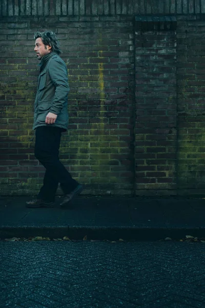 Homem de casaco verde andando no pavimento ao longo da parede de tijolo . — Fotografia de Stock