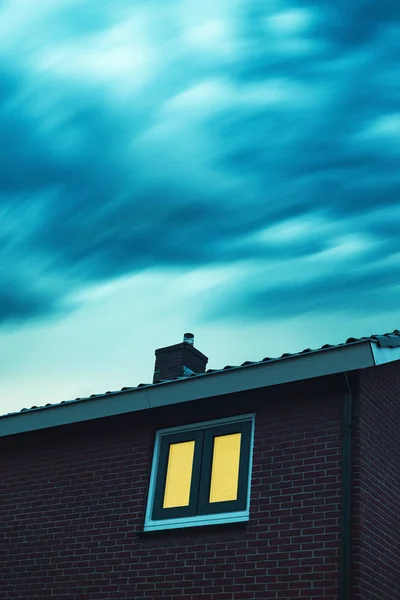 Straszny dom z oświetlonym oknem pod burzliwym niebem o zmierzchu. — Zdjęcie stockowe