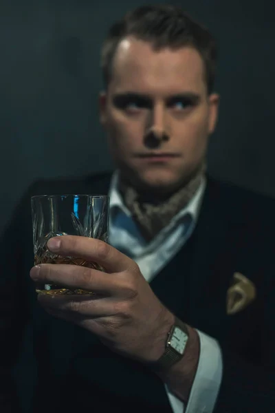 Hombre de moda retro en traje oscuro sostiene un vaso de whisky . — Foto de Stock